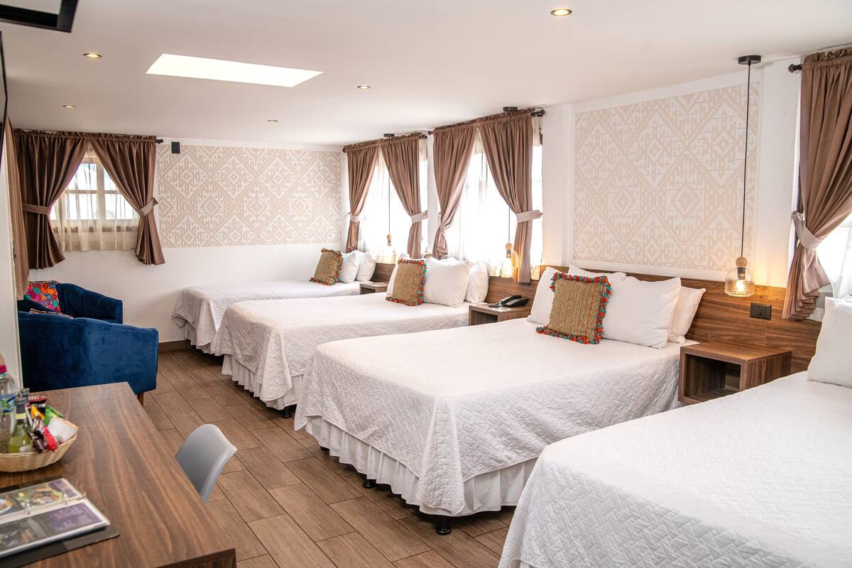 A hotel room with three single beds, a blue armchair, a wooden table with a basket, and patterned wallpaper.
