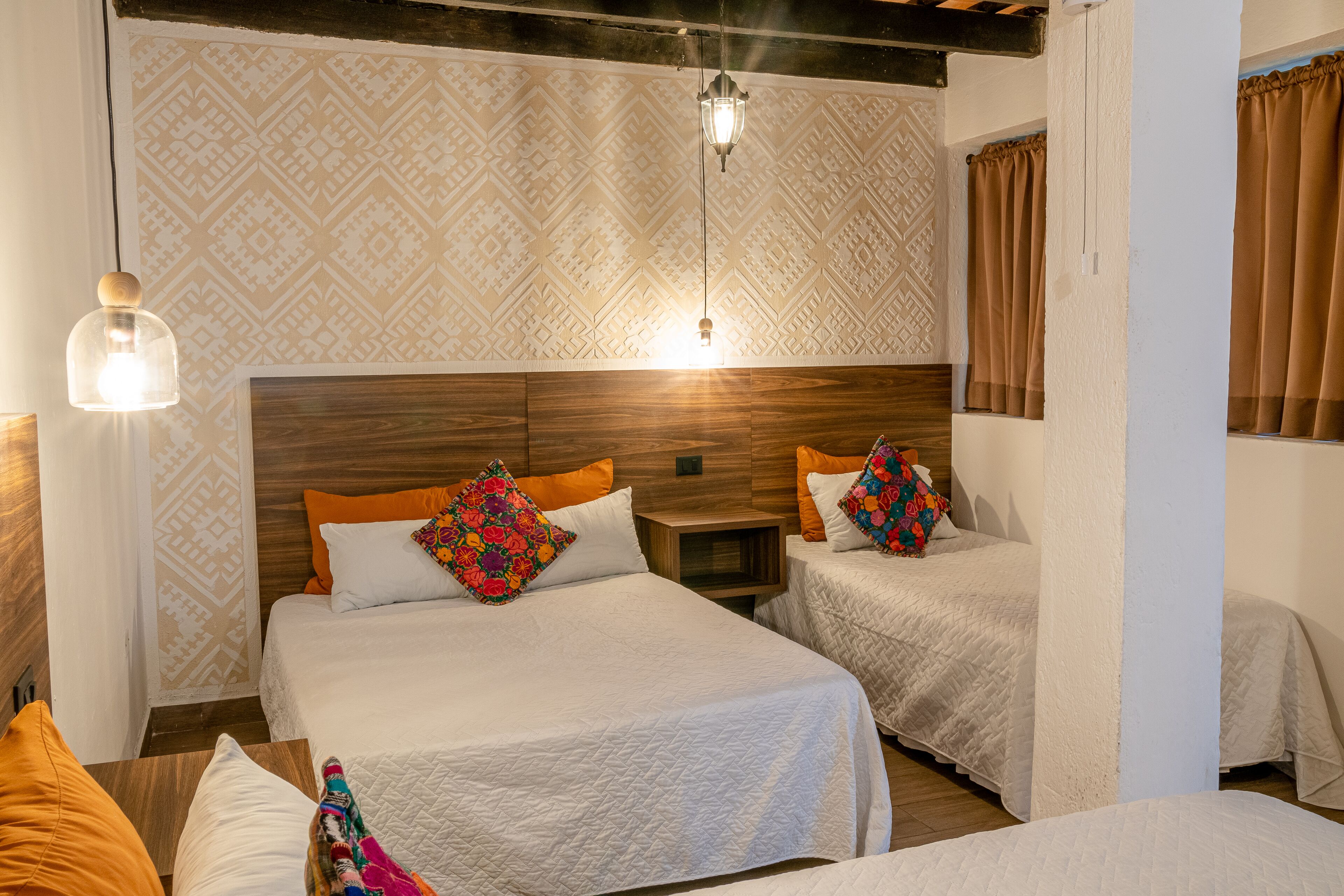 Una habitación con dos camas, cada una con ropa blanca y almohadas coloridas, un cabecero de madera, papel tapiz estampado y una lámpara colgante.