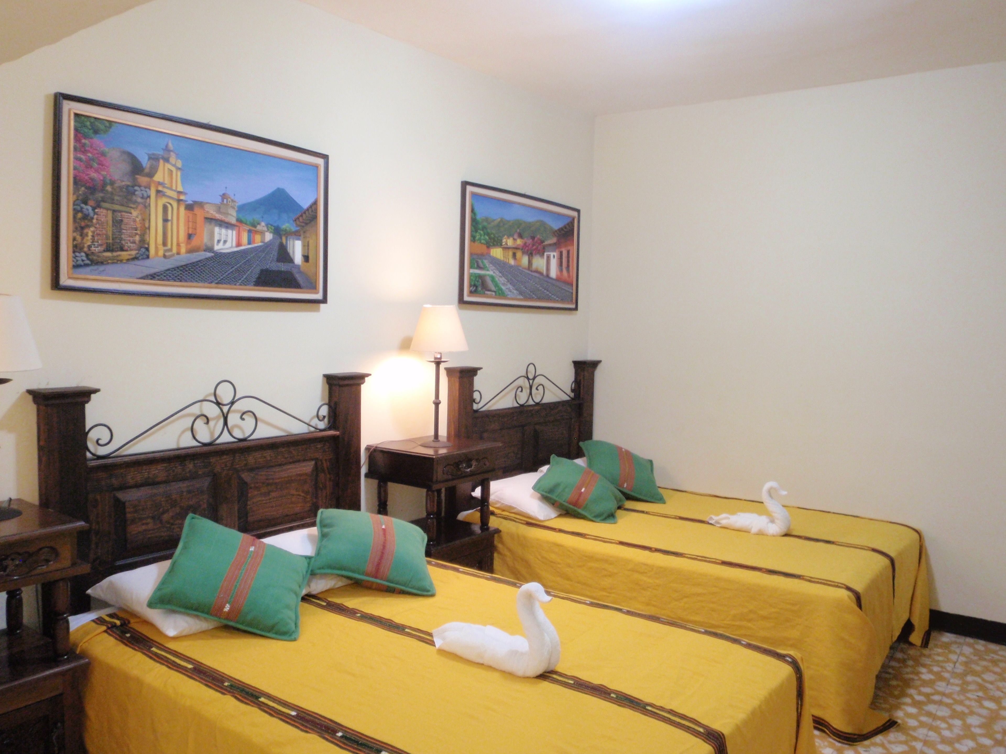 Une chambre d’hôtel avec deux lits, chacun recouvert d’une couvre-lit jaune et agrémenté d’oreillers verts, une tête de lit en bois, une table de chevet avec une lampe, et deux tableaux encadrés accrochés au mur.