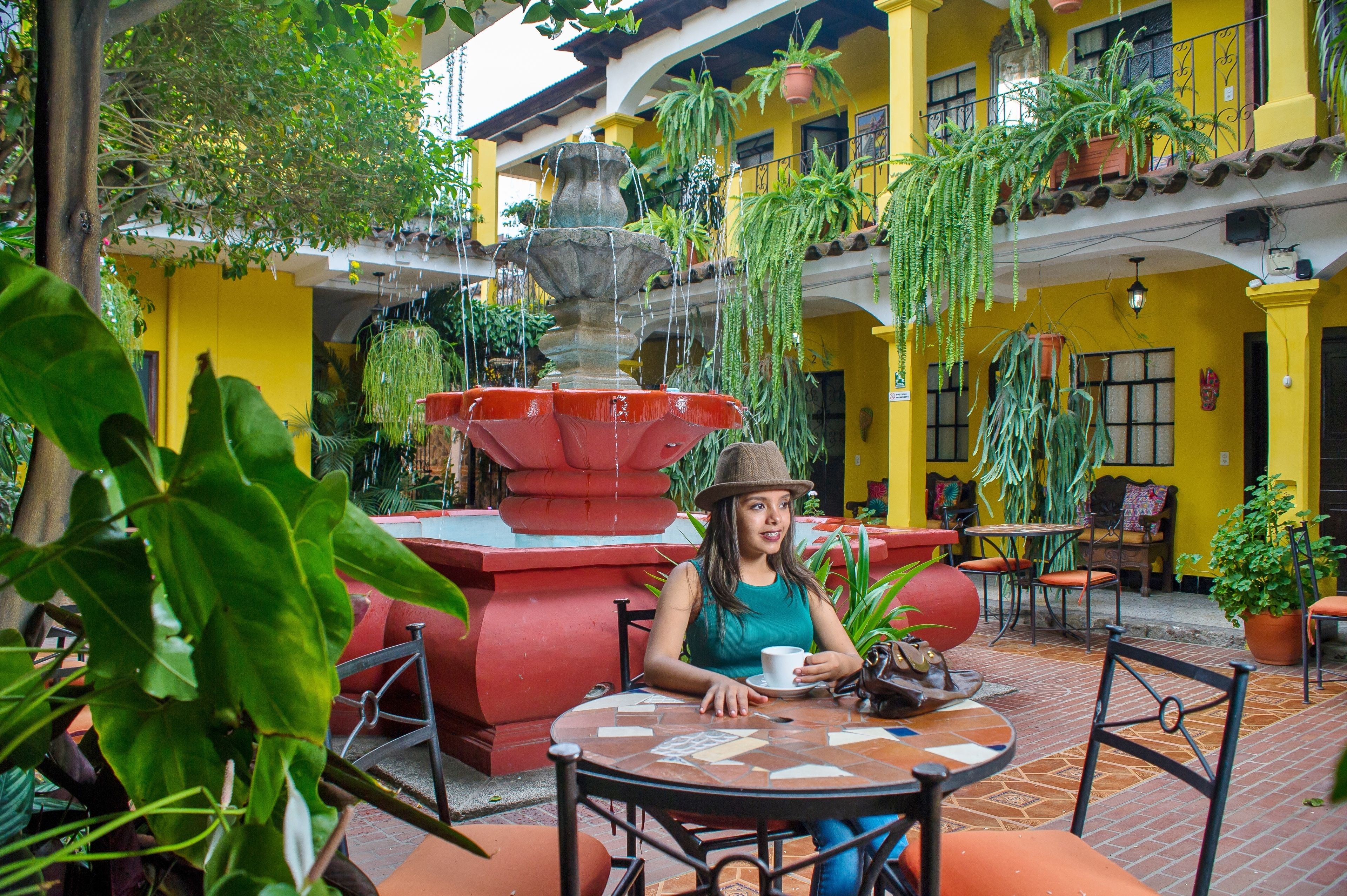 Una mujer sentada a una mesa con una taza, rodeada de macetas y una fuente en un patio con paredes amarillas y suelo de baldosas.