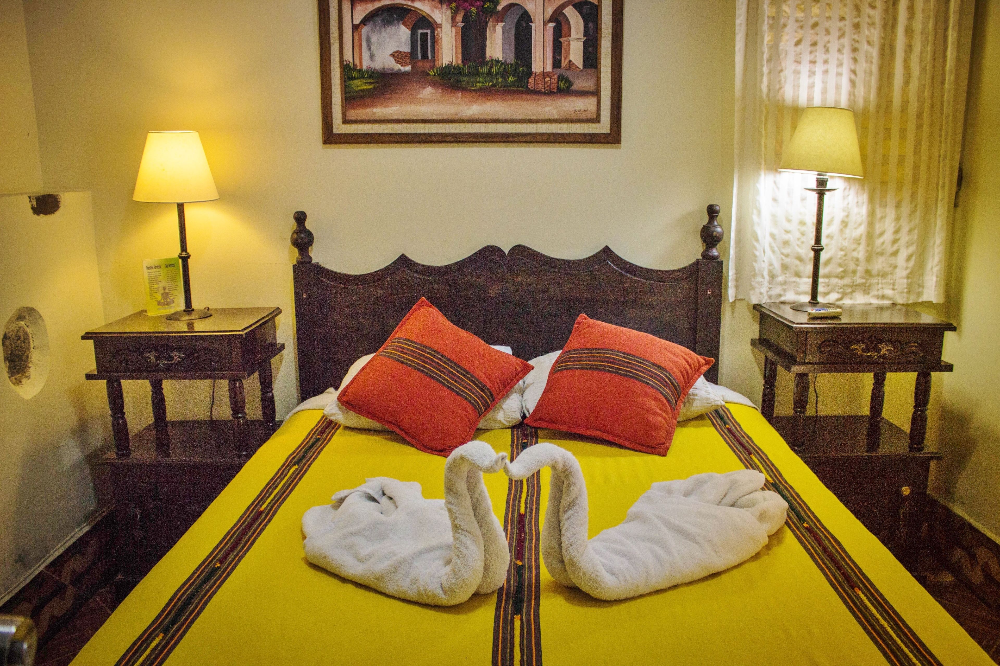 A hotel room with a bed, two red pillows, two white towels arranged in swan shapes, a painting on the wall, and two bedside lamps.