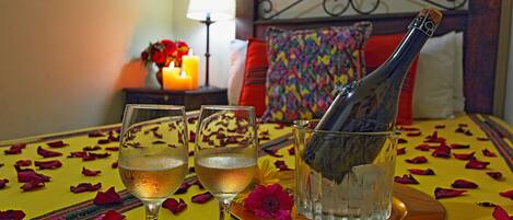 Un lit agrémenté d’une tête de lit décorative, une table de chevet avec une lampe, et une table sur laquelle se trouvent une bouteille de champagne, des verres et des pétales de rose.