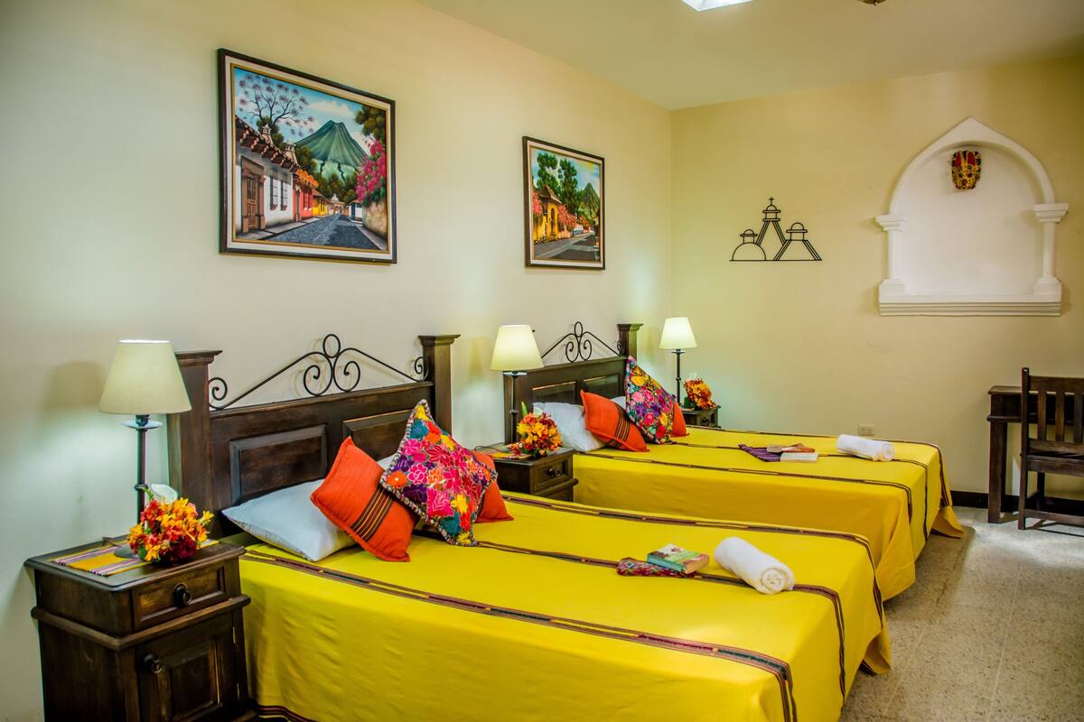 Habitación de hotel con cuatro camas, cada una con colcha amarilla y almohadas coloridas. Hay lámparas fijadas a la pared a ambos lados de las camas, y una pequeña mesa con lámpara y flores a la izquierda. La habitación tiene un diseño sencillo y limpio, con un pequeño arco en la pared derecha.