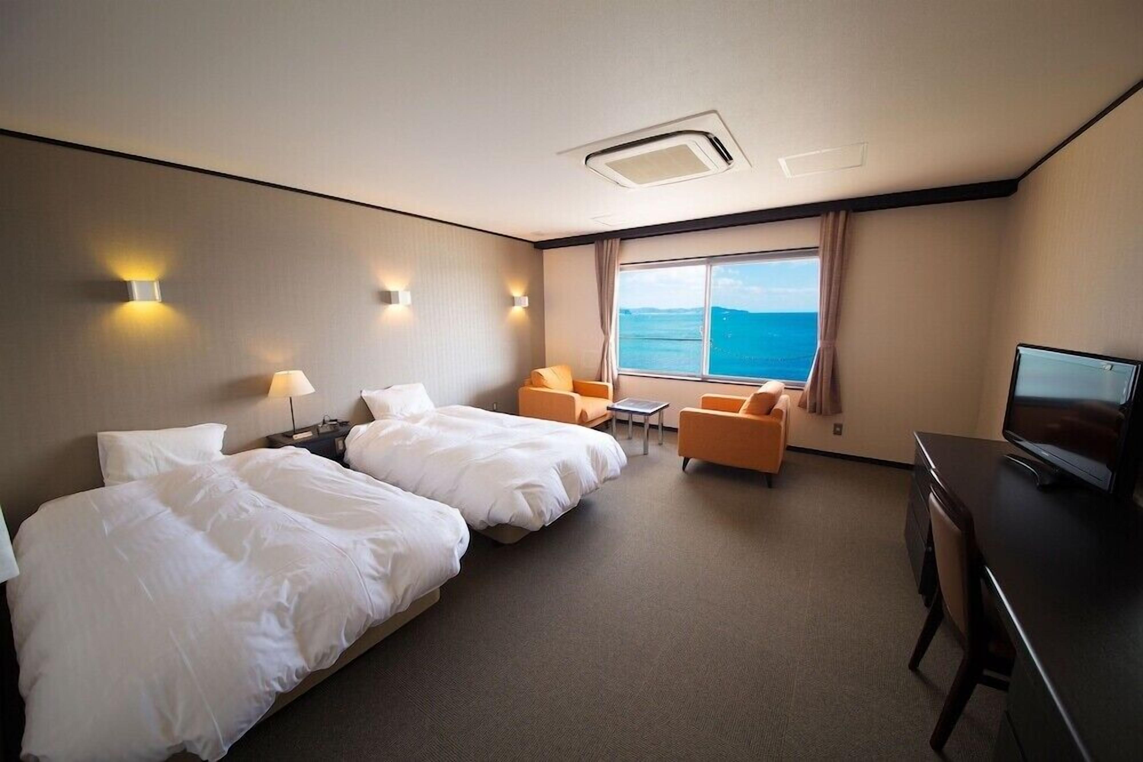 A hotel room with two beds, a desk, a TV, and a view of the sea.