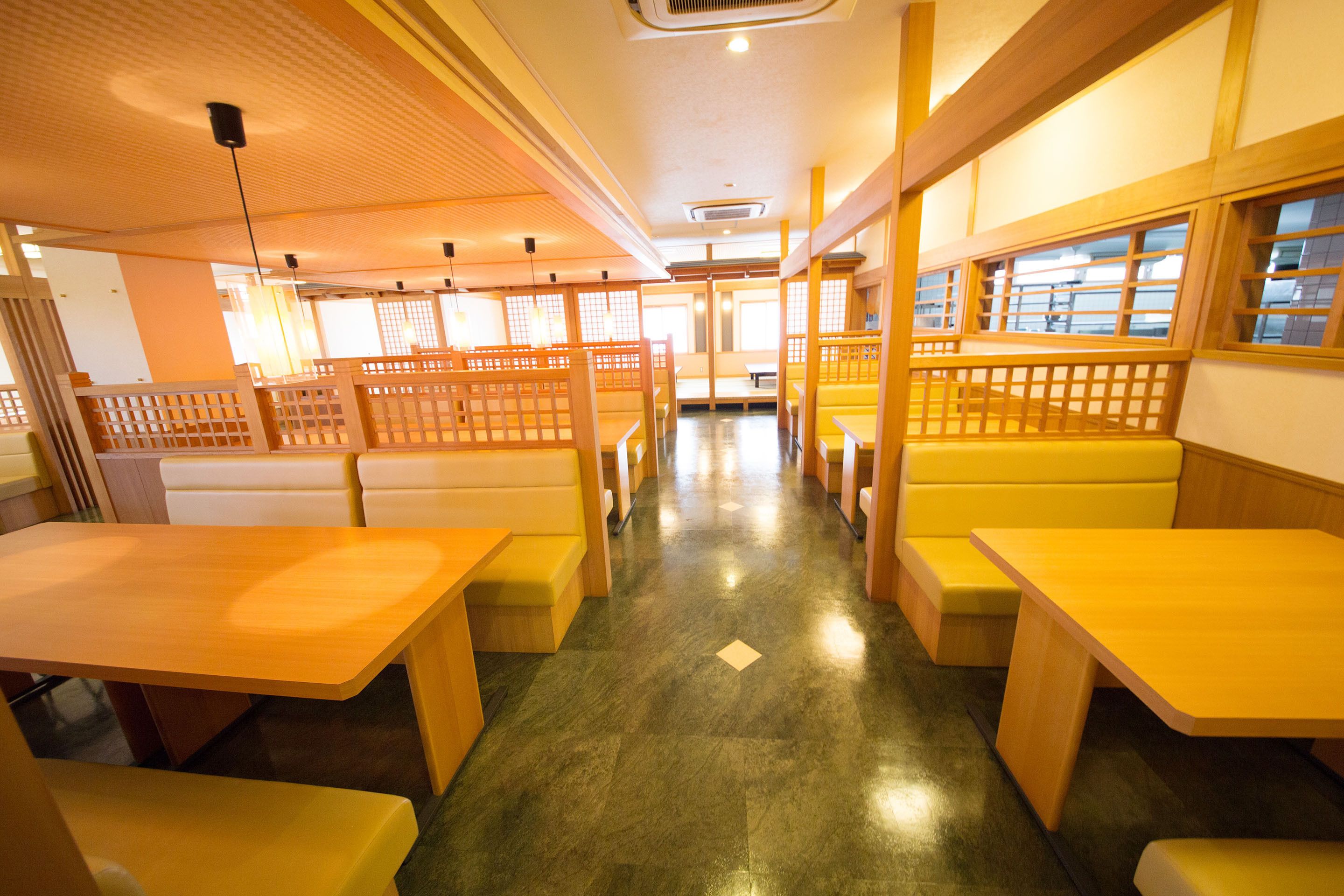 L’intérieur d’un restaurant présente des banquettes et des tables en bois, un sol carrelé et un plafond haut avec des luminaires encastrés.