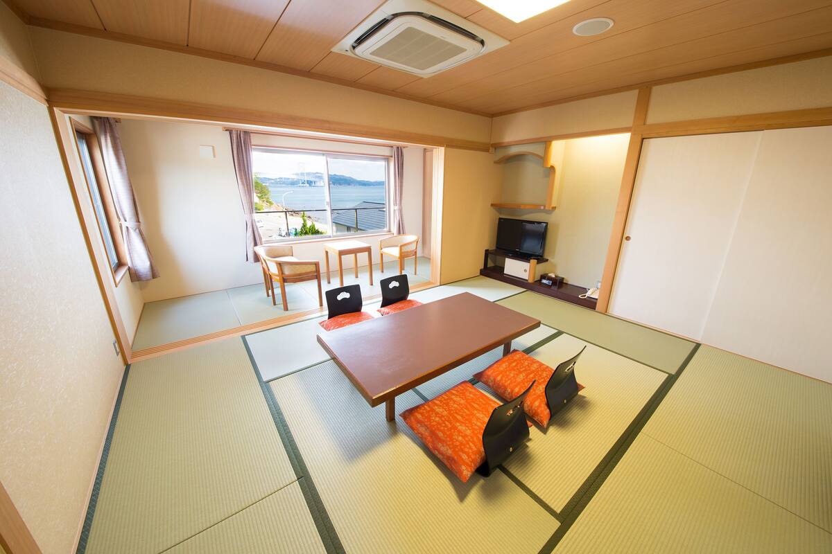 A traditional Japanese room with tatami flooring, a low table with cushions, a television, and a view of the sea through large windows.