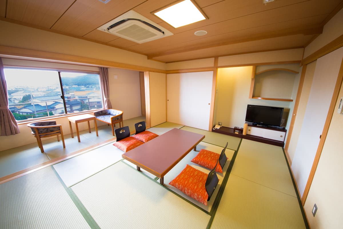 A traditional Japanese room with tatami flooring, a low table with cushions, a television, and a view of the city.
