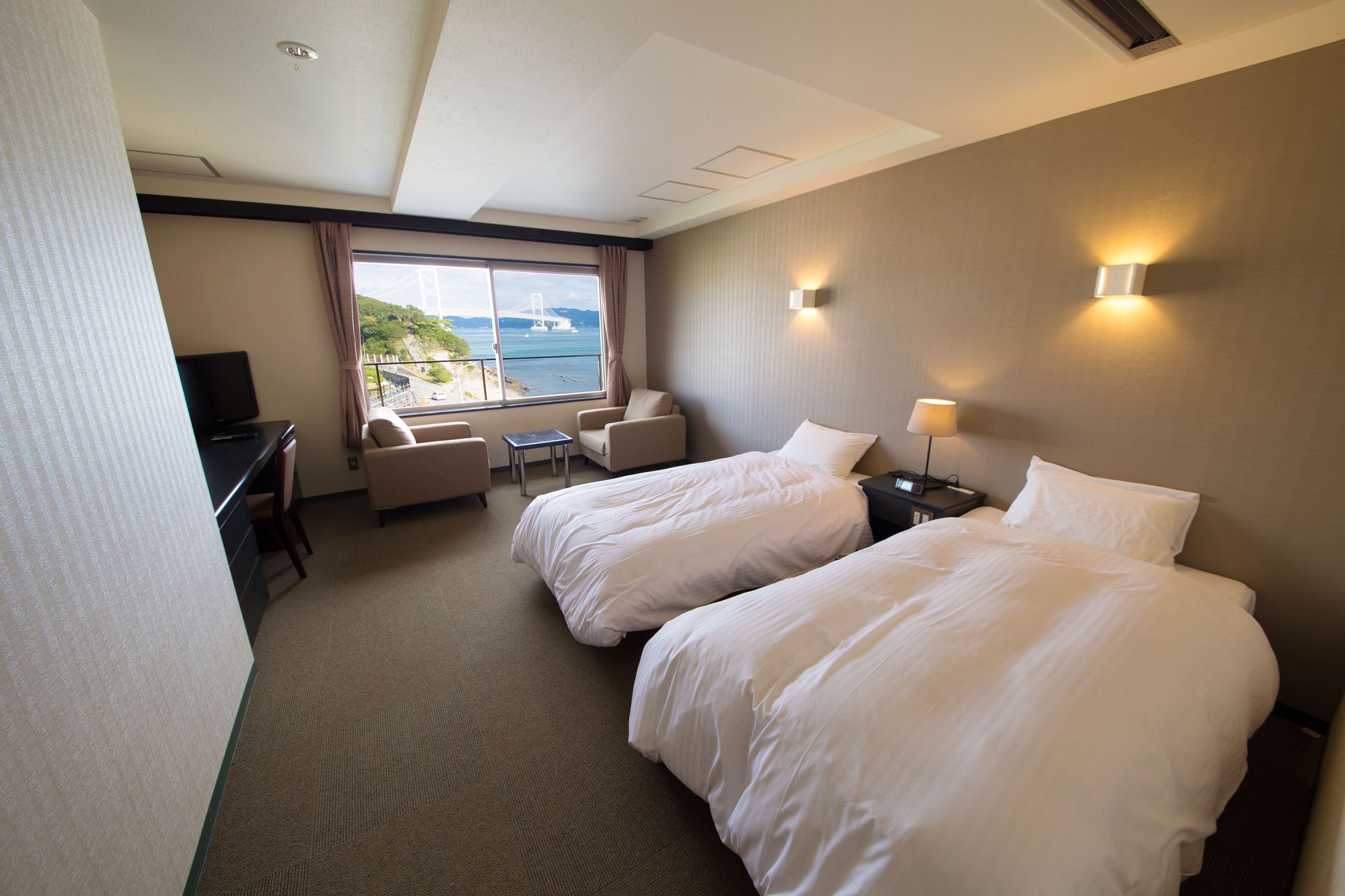 A hotel room with two beds, a desk with a chair, a TV, and a window with a view of a bridge and water.