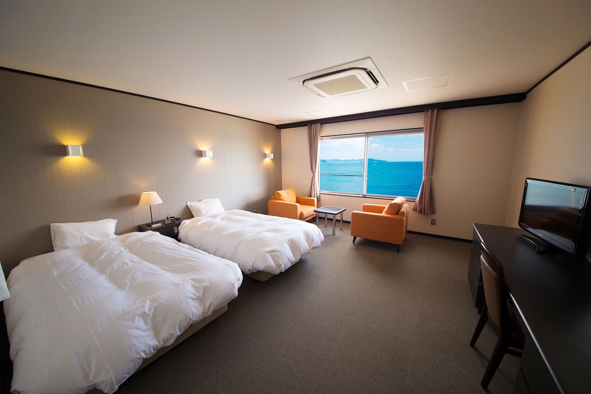 A hotel room with two beds, a desk, a TV, and a view of the sea.