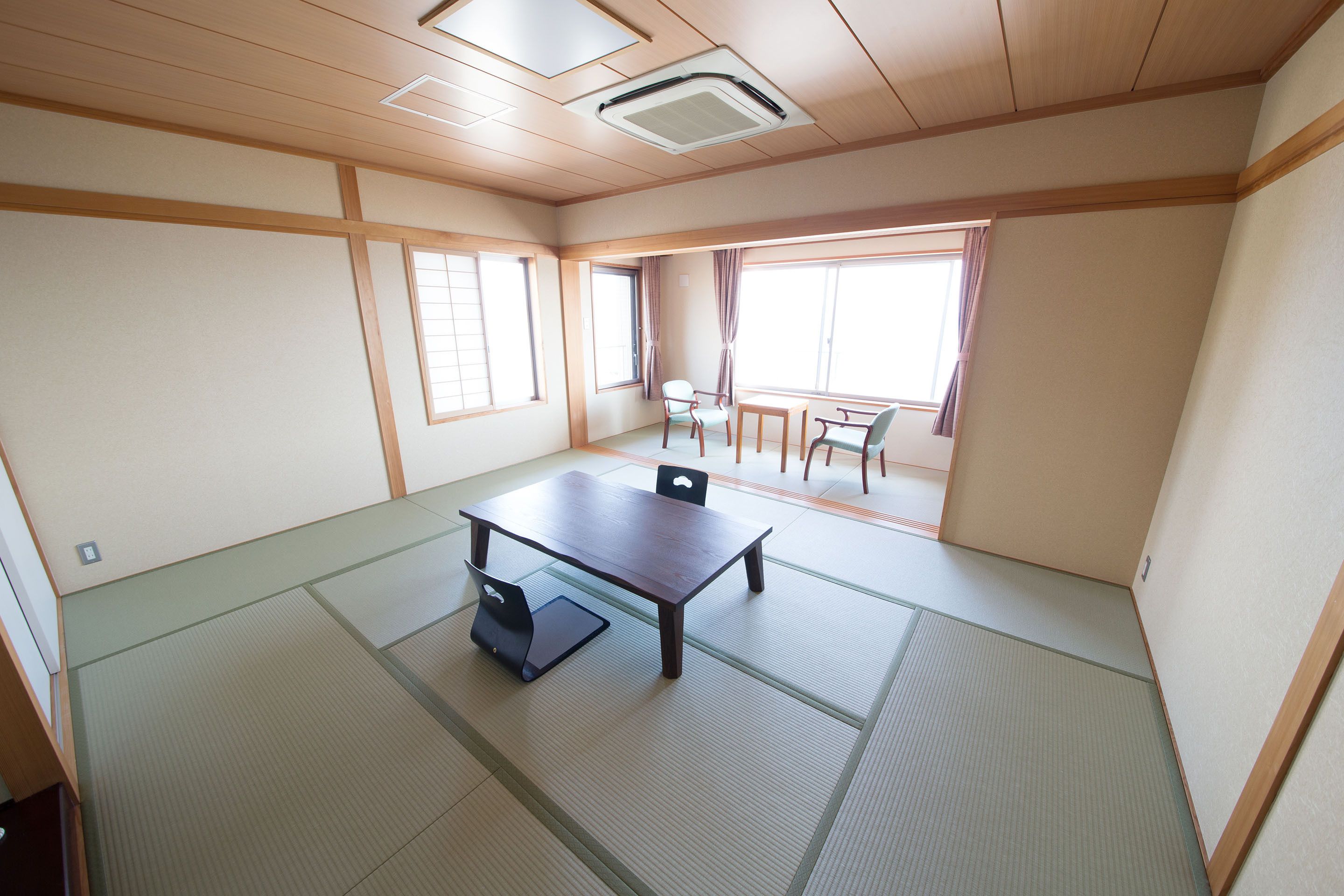 Een kamer met tatami vloerbedekking, een lage tafel met stoelen, en grote ramen die natuurlijk licht binnenlaten.