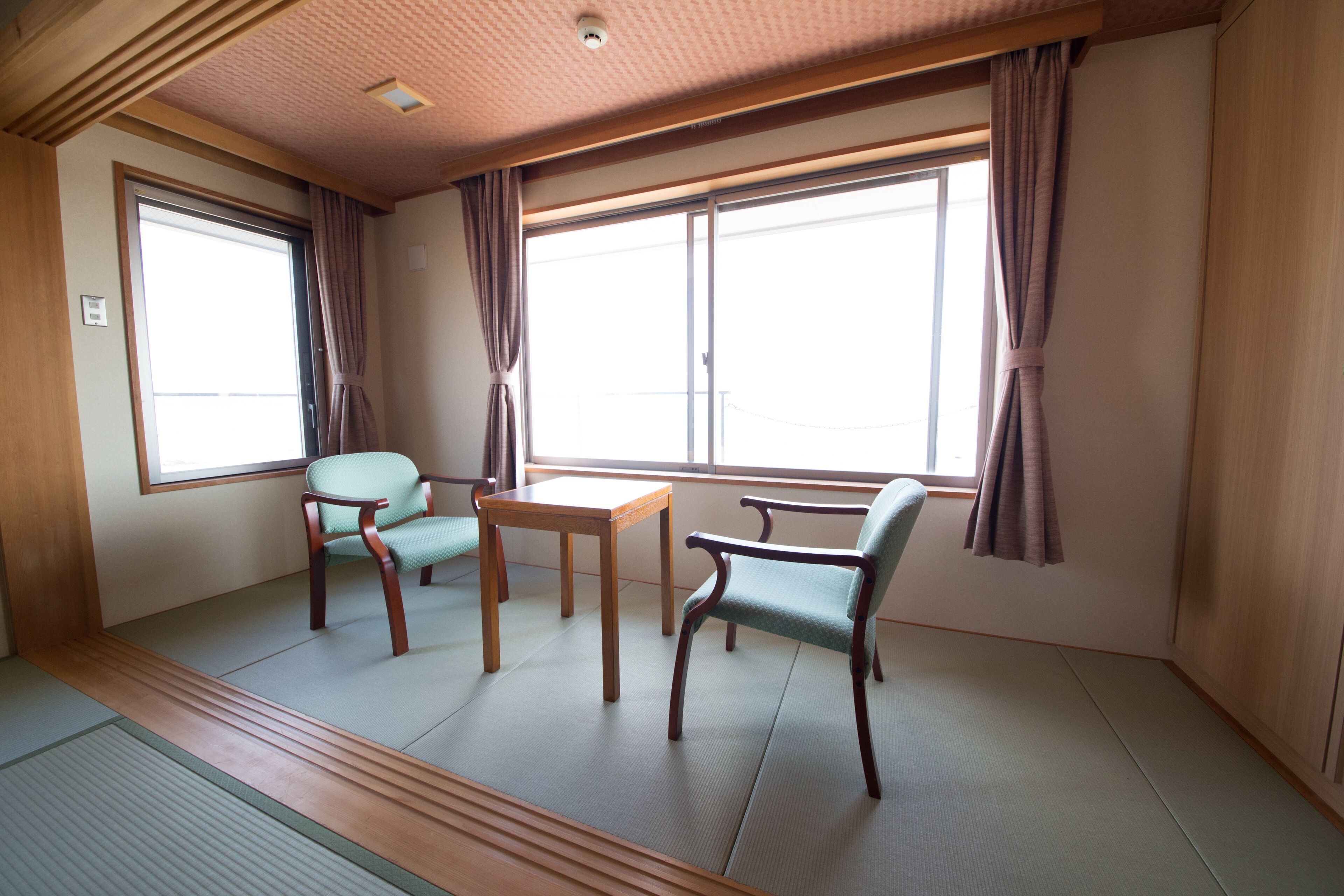 A room with tatami flooring, a wooden table, two chairs, and large windows with curtains.