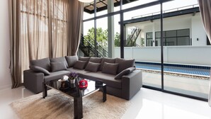 Un salon moderne avec un canapé gris, une table basse en verre et une vue sur une piscine, visible à travers de grandes fenêtres.