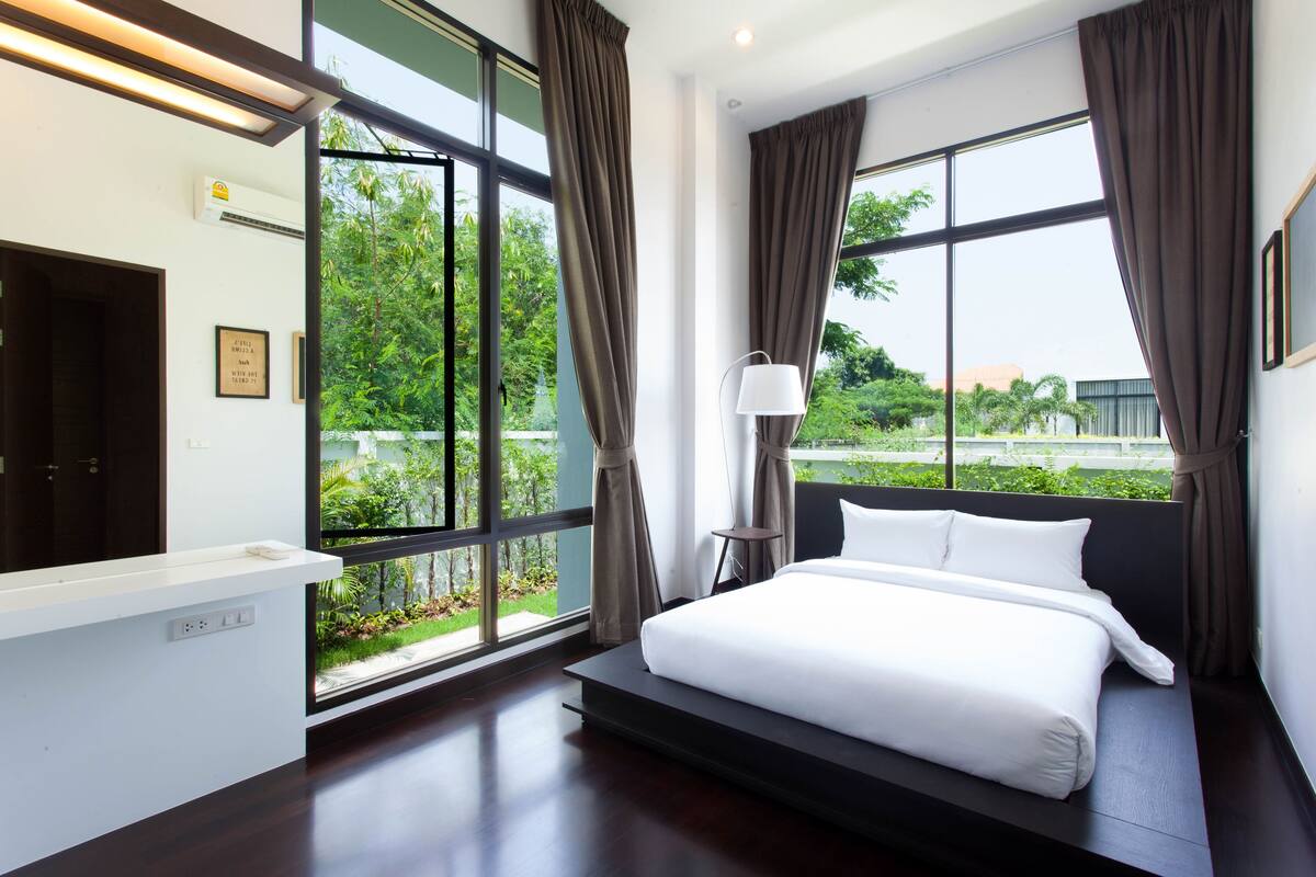 Habitación de hotel moderna con una cama grande, una mesita de noche, una lámpara y vistas a zonas verdes a través de amplios ventanales.