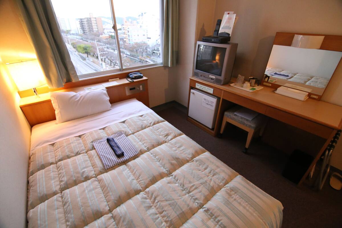 A hotel room with a bed, a desk with a television, a small refrigerator, and a window with a view of buildings.