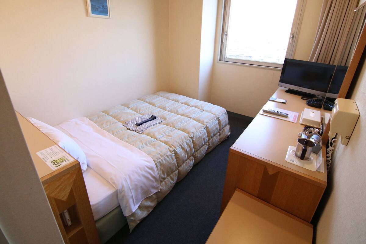 A hotel room with a bed, a desk with a computer, a TV, and a window with curtains.