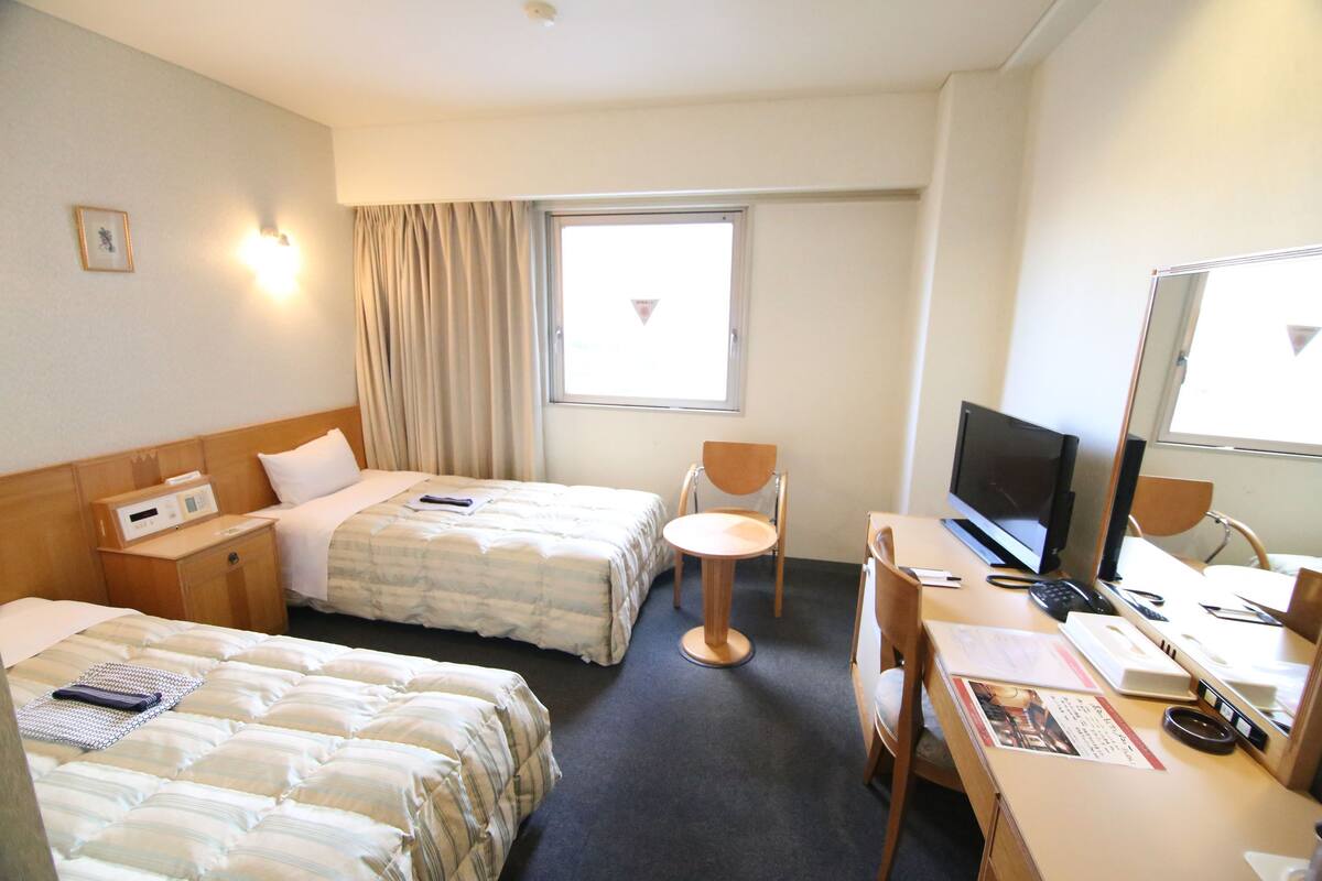 A hotel room with two beds, a desk with a computer, a chair, and a window with curtains.