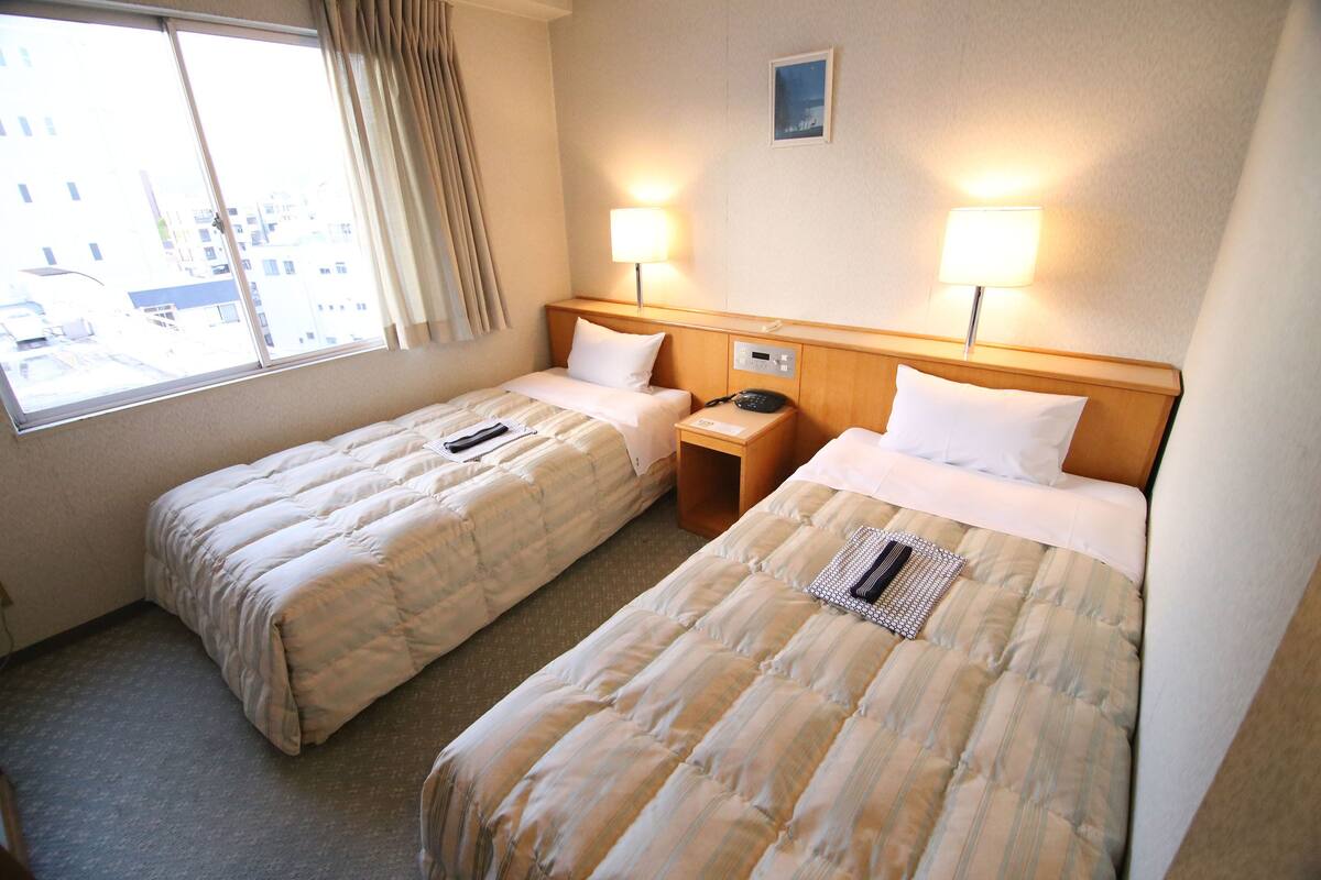 A hotel room with two single beds, a window with curtains, bedside lamps, a small table, and a wall-mounted phone.