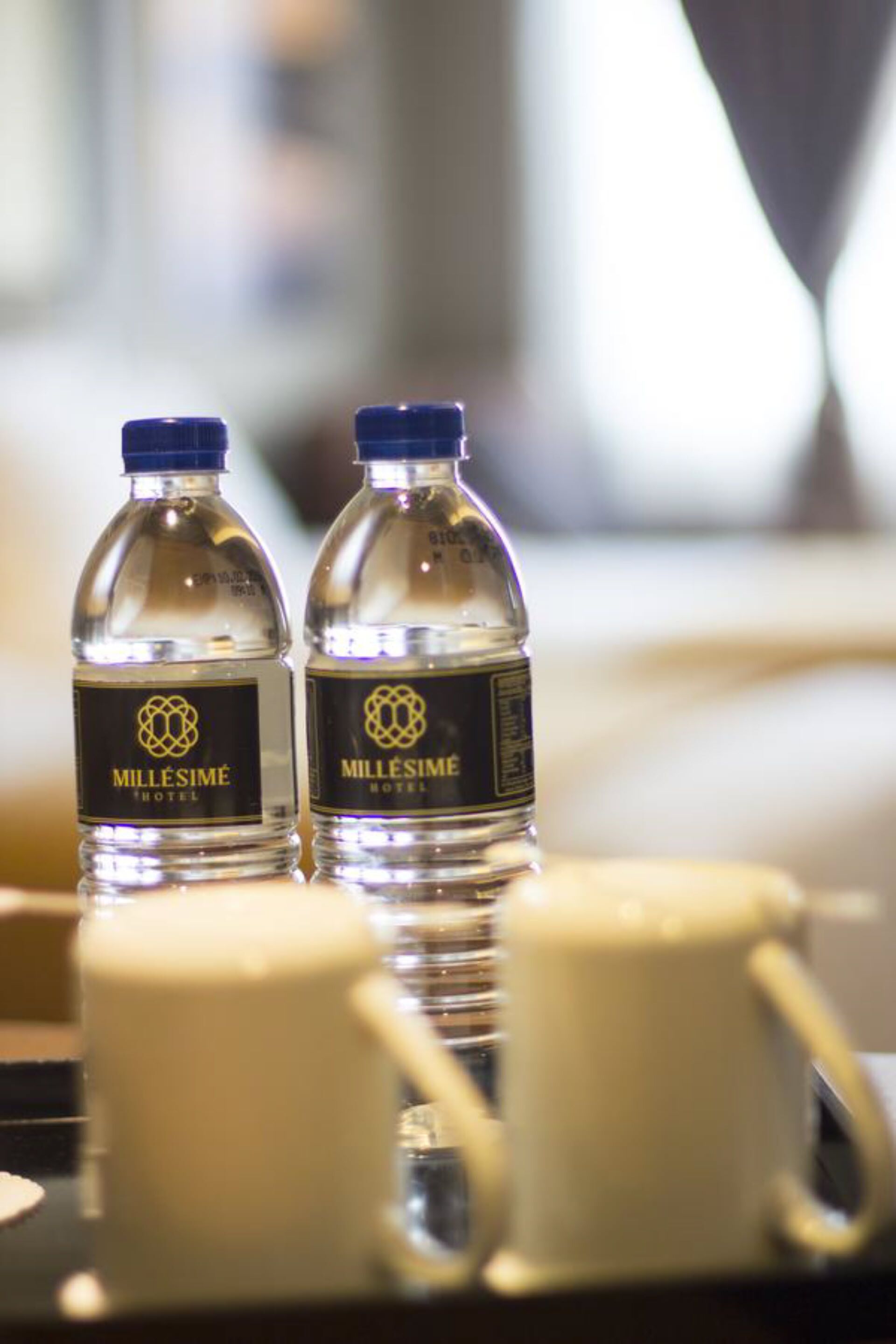 Two water bottles labeled 'Millésime Hotel' on a tray with two white mugs.