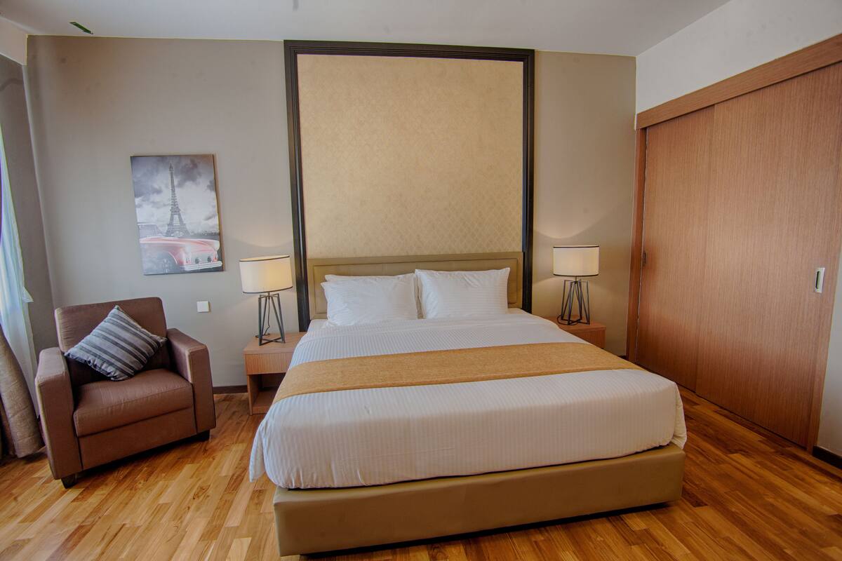 A hotel room with a large bed, a wooden floor, a brown armchair, a nightstand with lamps, and a door.