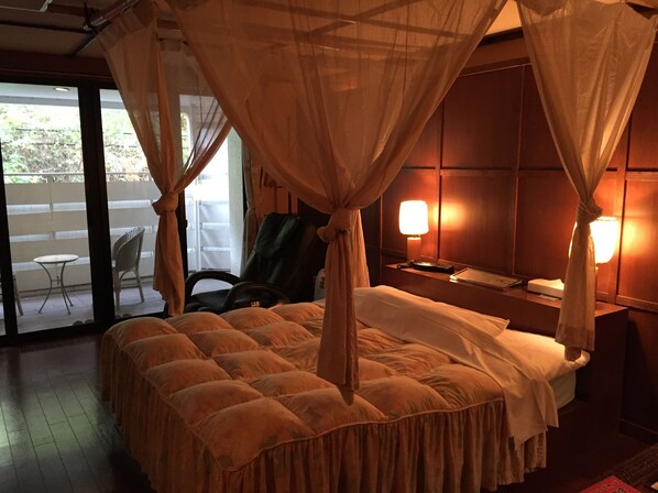 A hotel room with a four-poster bed, a desk with a lamp, a chair, and a view of the outdoors through a sliding door.