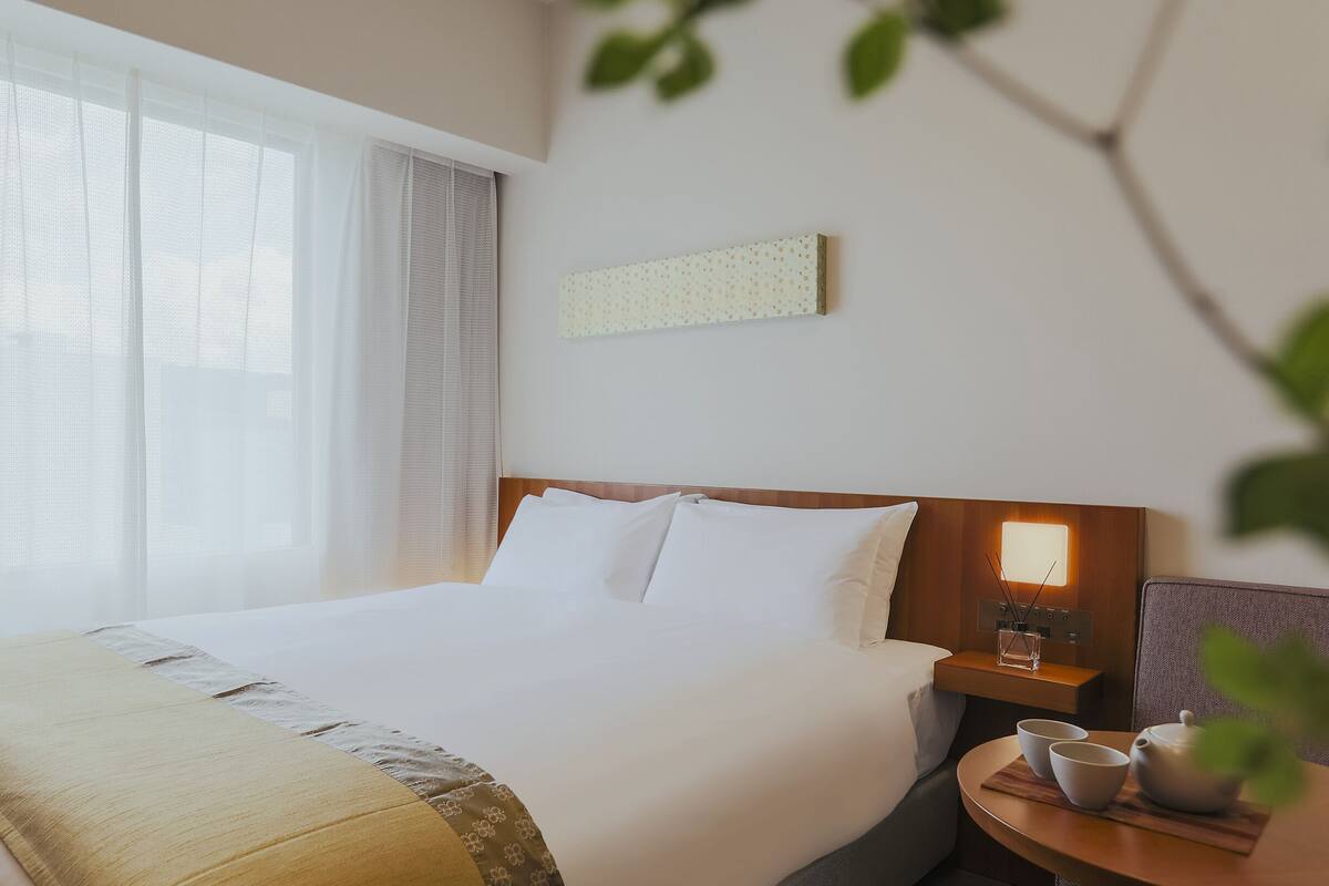 Hotellihuone, jossa on suuri sänky, yöpöytä lampun kanssa ja näkymä ulos ohuiden verhojen läpi.