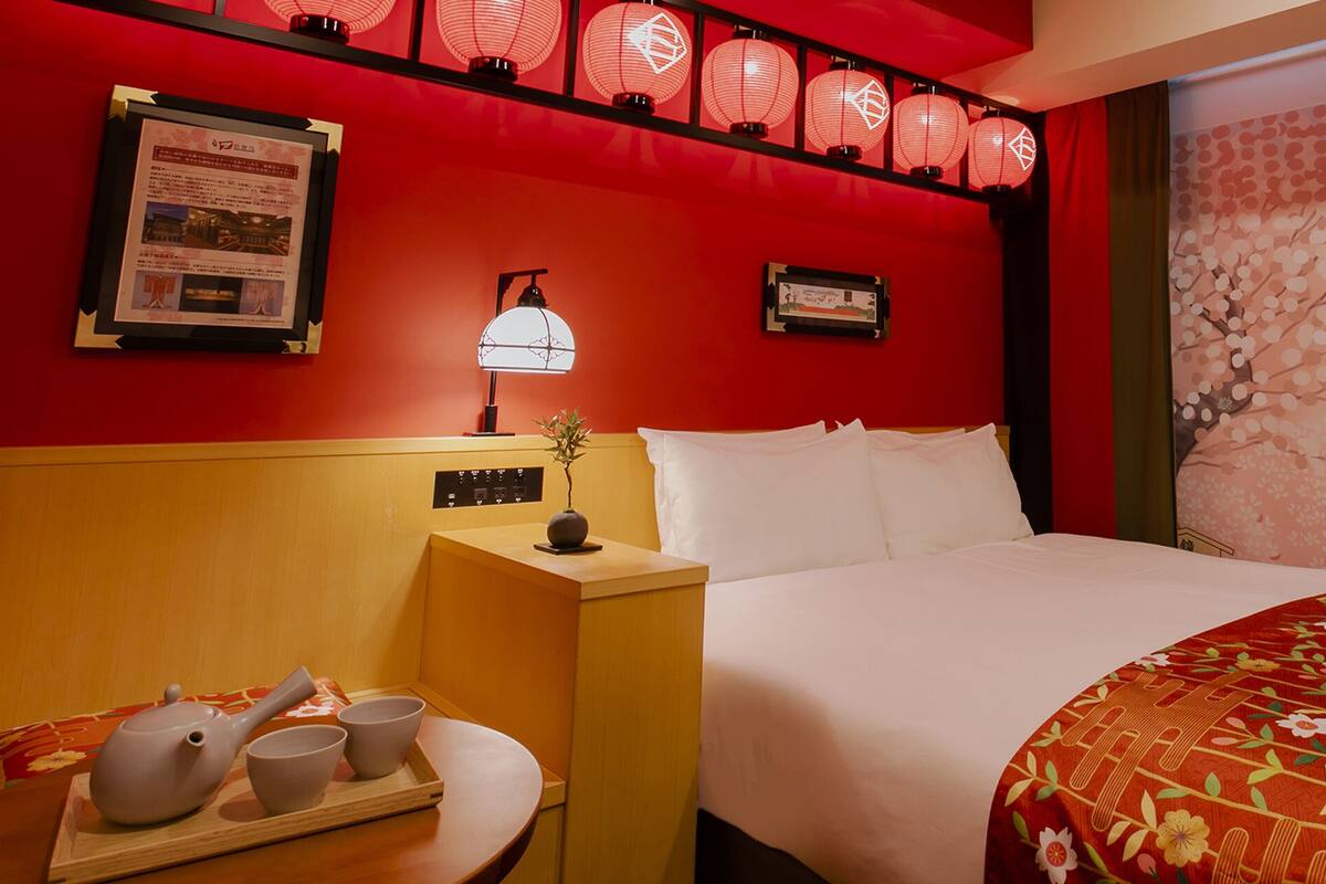 Hotellihuone, jossa on sänky, yöpöytä lampun kanssa, tarjoitinalusta teekannulla ja kupeilla sekä punainen seinä, jossa on lyhtyjä.