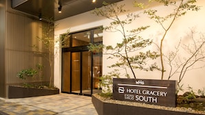 The entrance to Hotel Gracery Kyoto Sanjo South with a signboard, a tiled pathway, and a small garden area with plants.