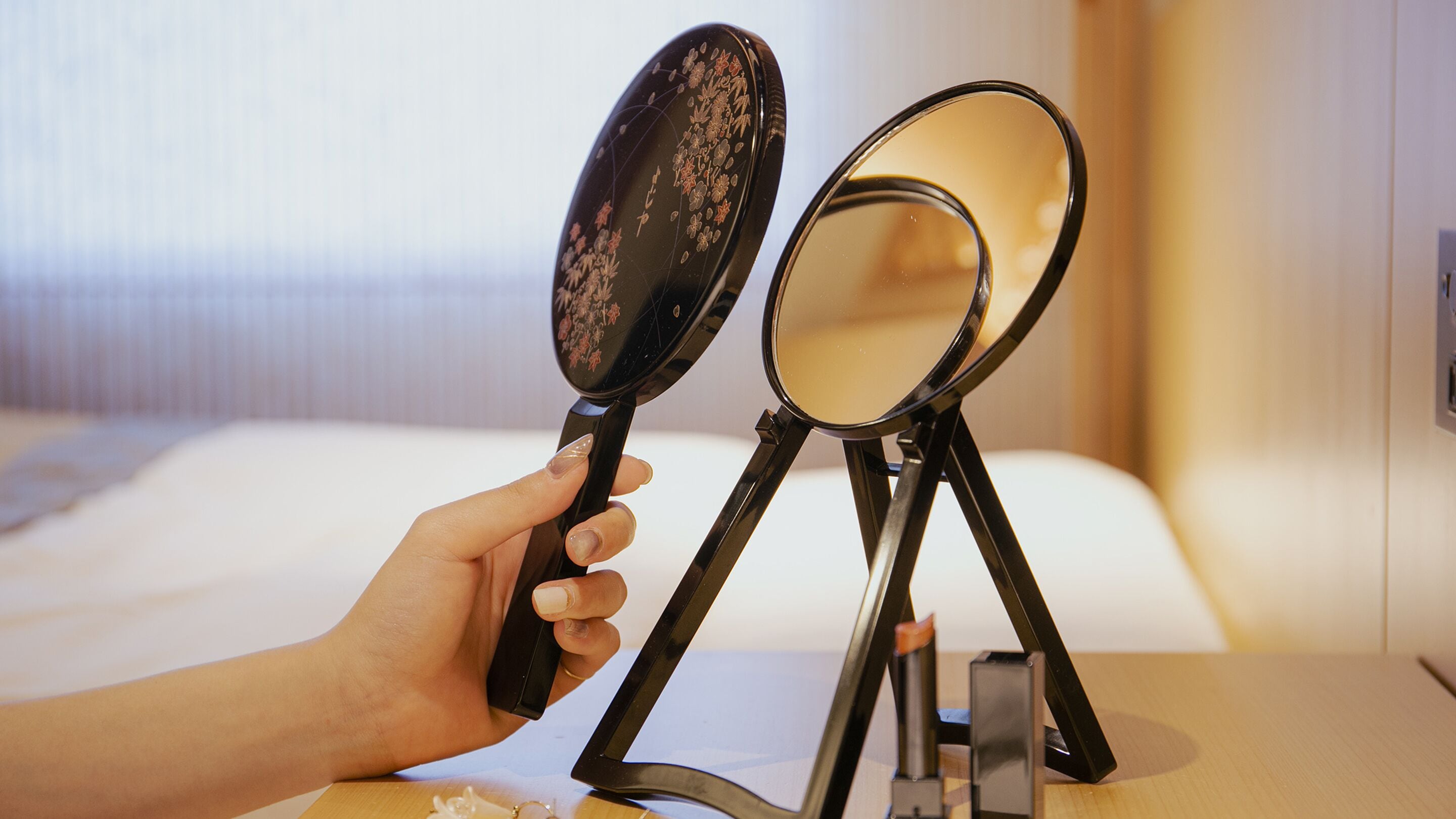 A hand holding a compact mirror with floral design, placed on a stand next to a larger mirror with a plain reflective surface.