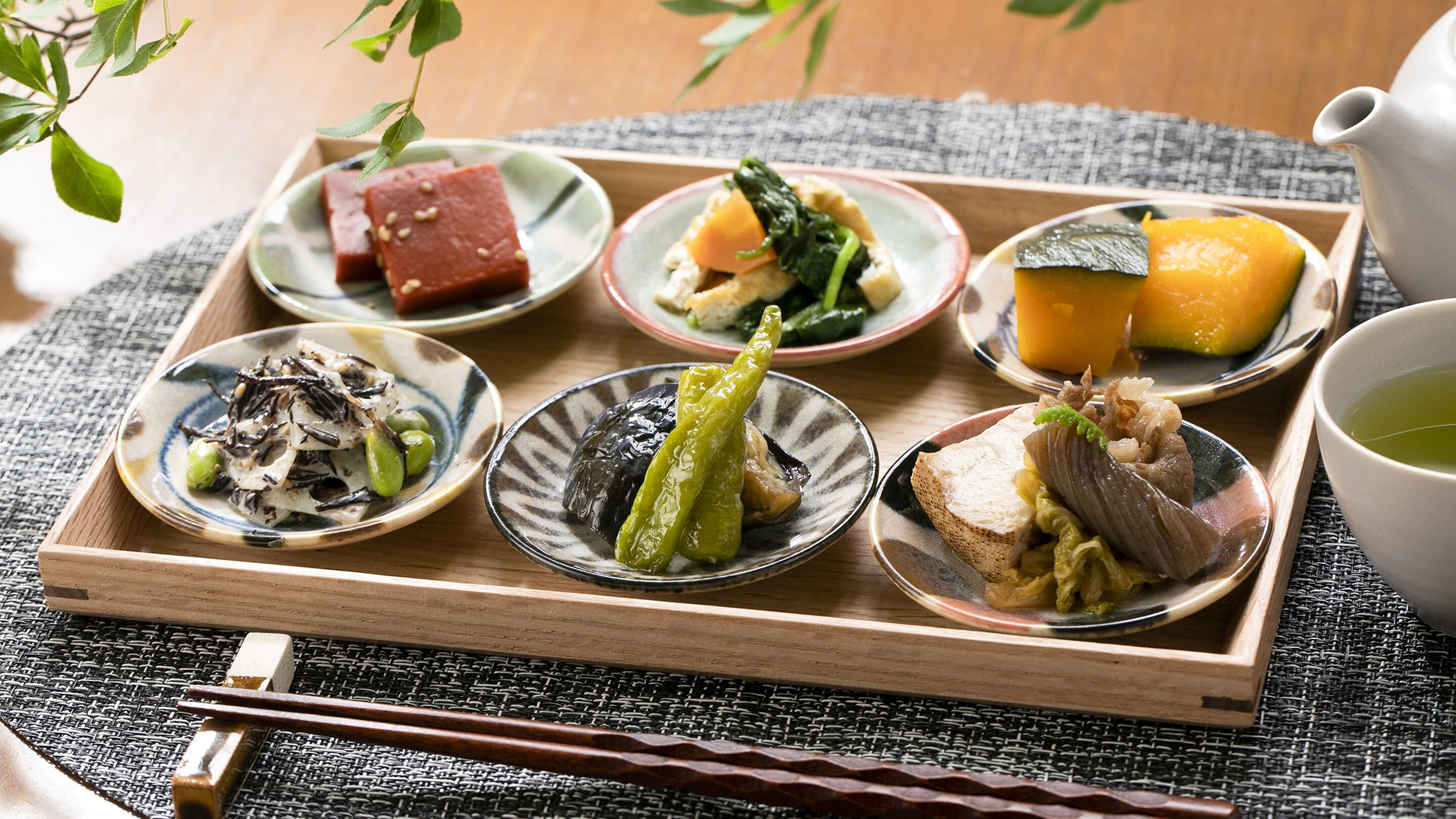 A wooden tray with various dishes, including tofu, vegetables, and meat, accompanied by chopsticks and a teapot.
