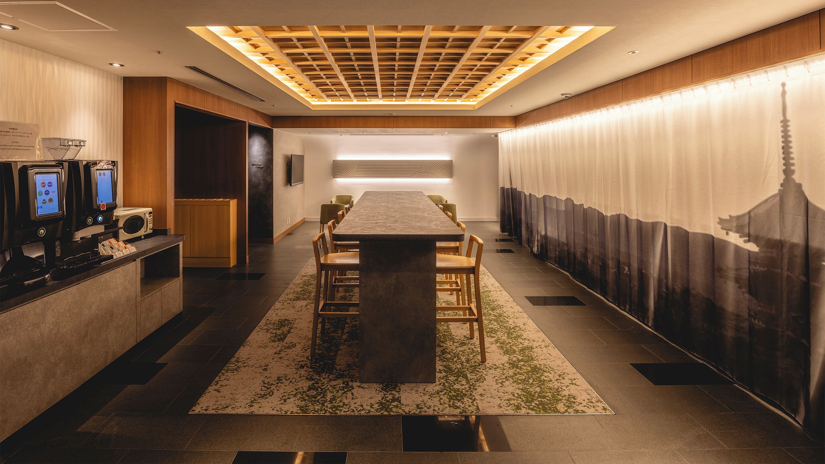 A hotel lobby with a long table, chairs, a reception desk with digital kiosks, a large wall mural, and a ceiling with a wooden panel design.
