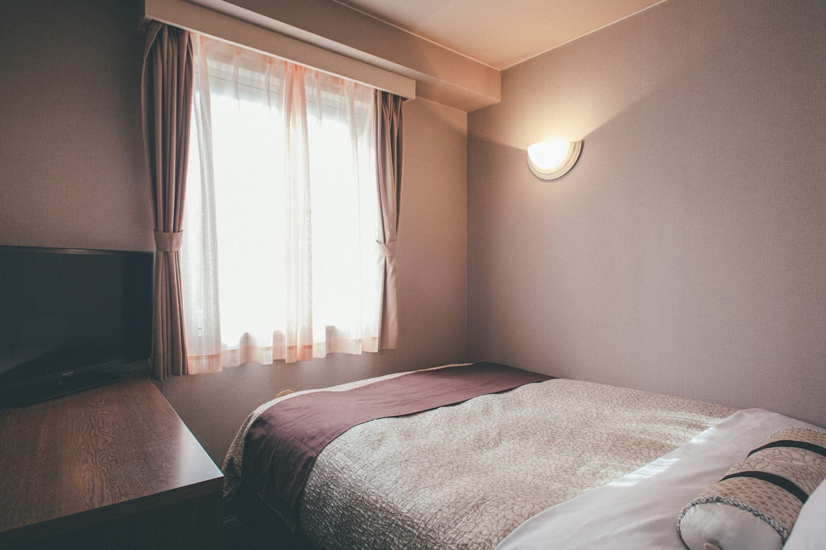 A hotel room with a bed, a TV, a window with curtains, and a wall-mounted light.