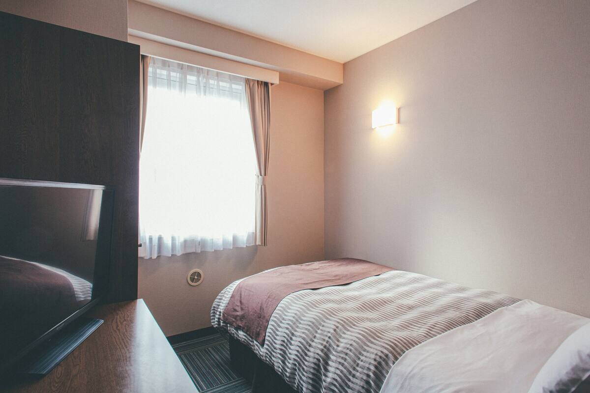 A hotel room with a bed, a television, a window with curtains, and a wall-mounted light.