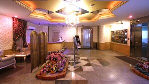Um hall de hotel com uma mesa de recepção, um candelabro, um painel fixado na parede e um piso estampado.