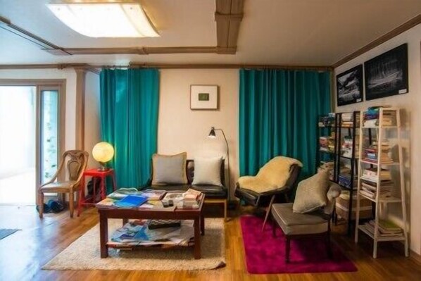 A living room with a wooden floor, a coffee table with magazines, a bookshelf, and teal curtains. - Pedro's House - Hostel (Gwangju)