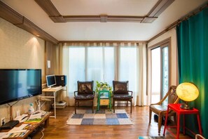 A hotel room with a large flat-screen TV, a desk with a computer, two leather chairs, a small table with a plant, a red lamp, and a checkered rug. - Pedro's House - Hostel (Gwangju)