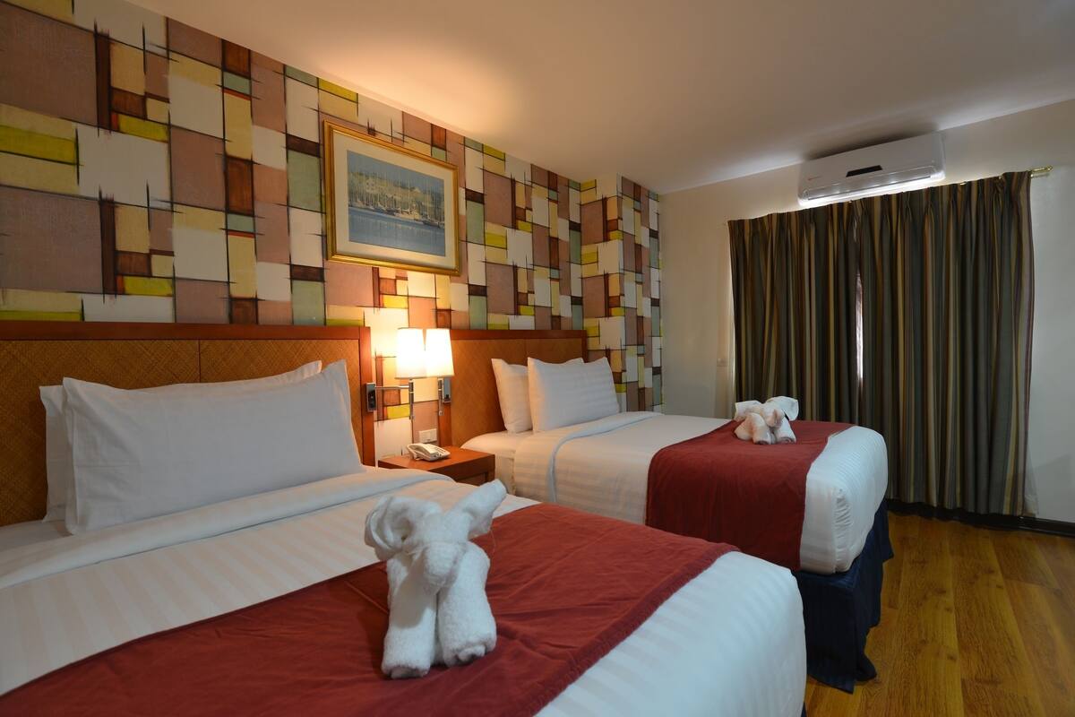 A hotel room with two beds, a patterned wall, a painting, and an air conditioning unit.