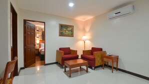 Una sala de estar con una mesa de centro de madera, dos sillones rojos, una pequeña mesa auxiliar de madera, una lámpara y un cuadro en la pared.