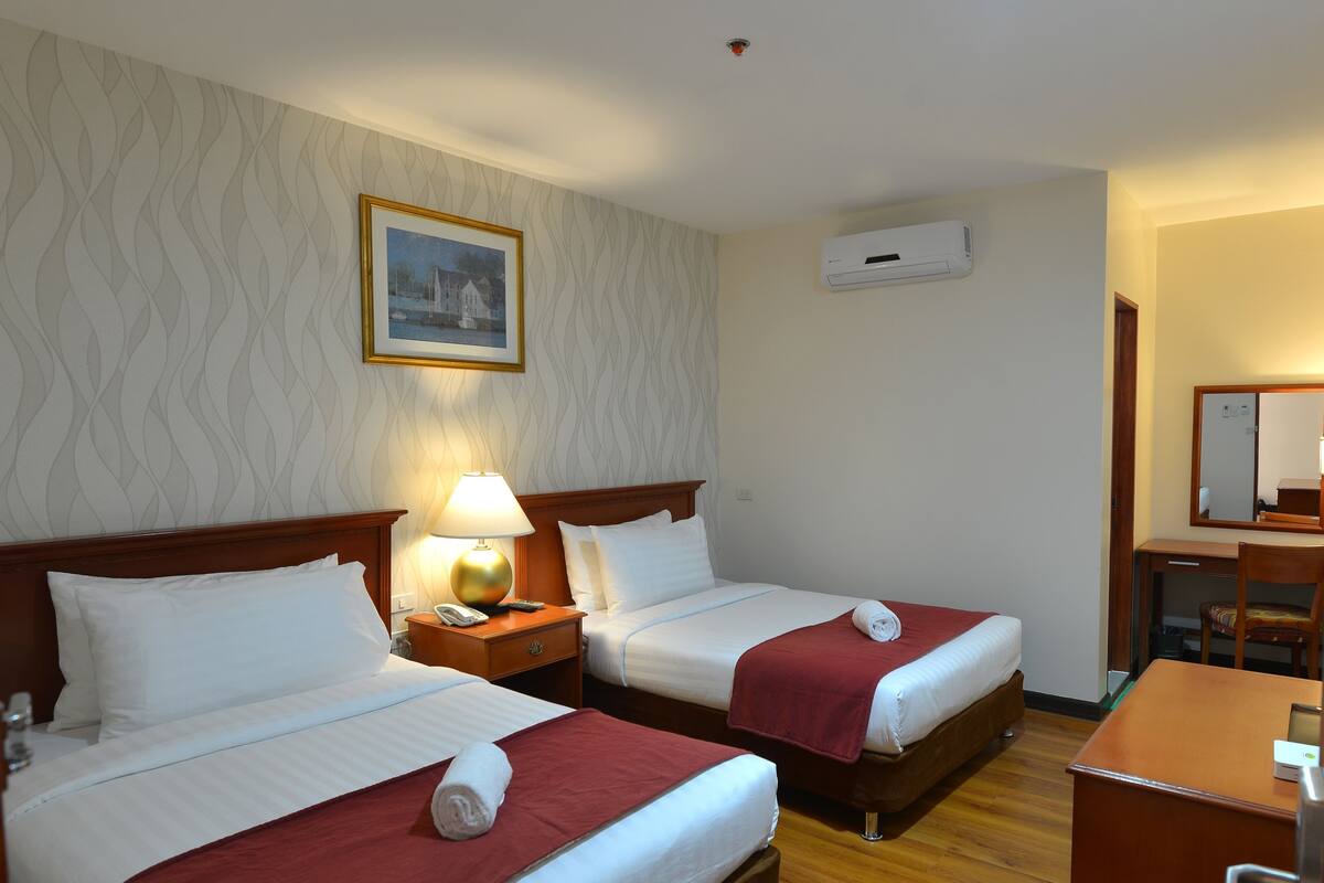A hotel room with two beds, a wooden desk, a lamp, and a painting on the wall.