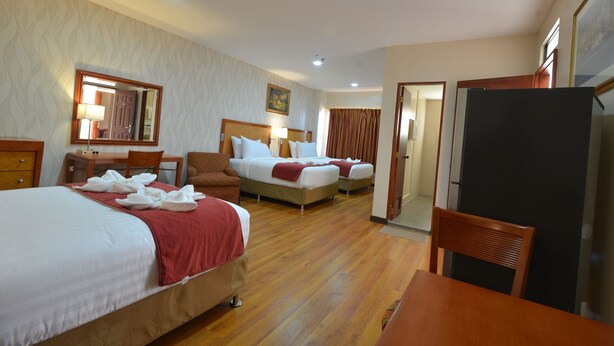 Habitación de hotel con tres camas, un escritorio, una silla, un refrigerador y una puerta que da a otra habitación.