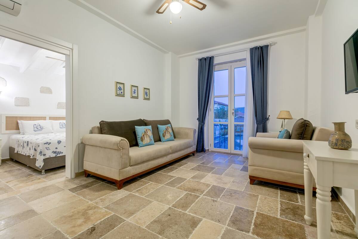 Una sala de estar con un sofá color crema, un sillón blanco, una mesita y una cama con colcha estampada.