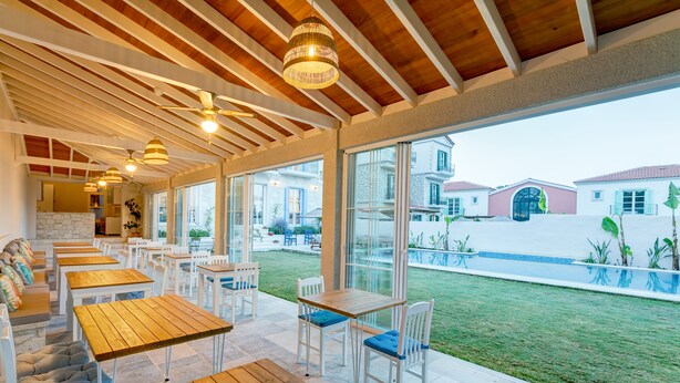 Un patio al aire libre con mesas de madera y sillas blancas, grandes ventanales que ofrecen vistas a un jardín trasero y casas vecinas.