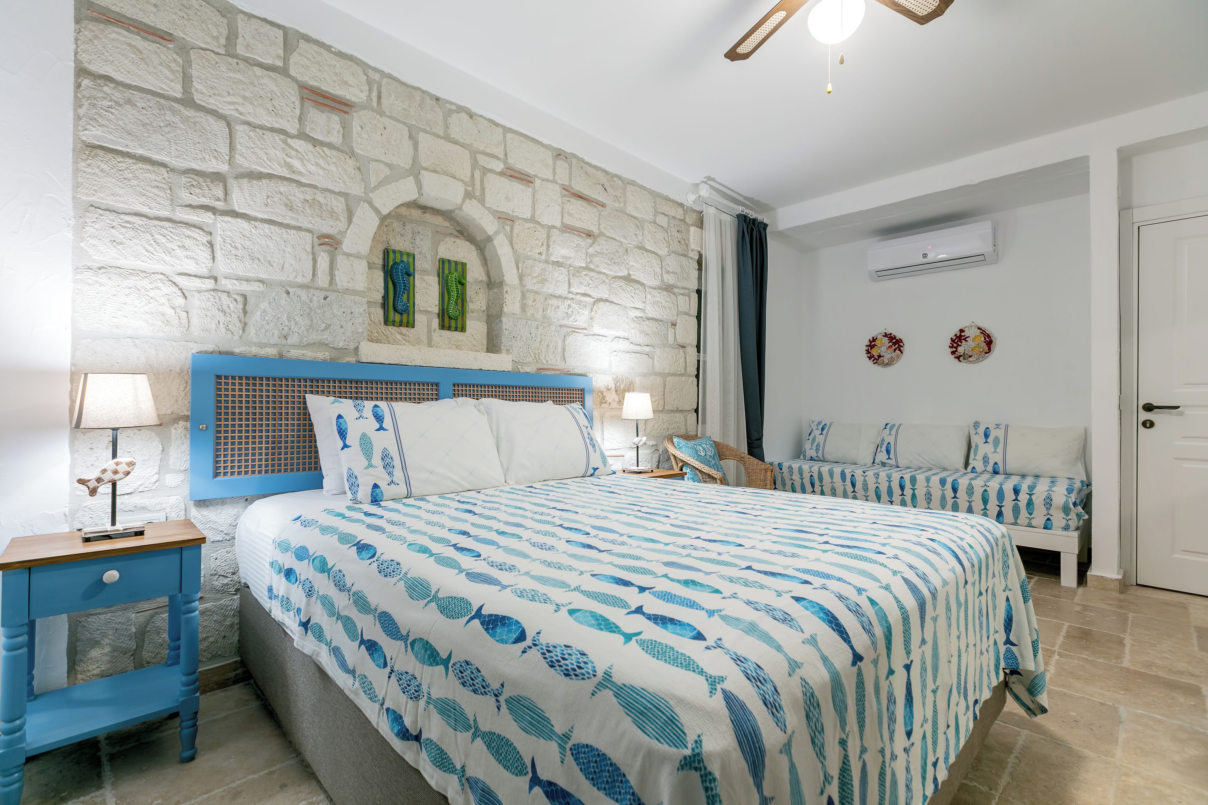 Un dormitorio con pared de piedra, cama con colcha de diseño azul y blanco con motivos de peces, dos mesitas de noche con lámparas y una pequeña zona de estar con un banco y cojines.