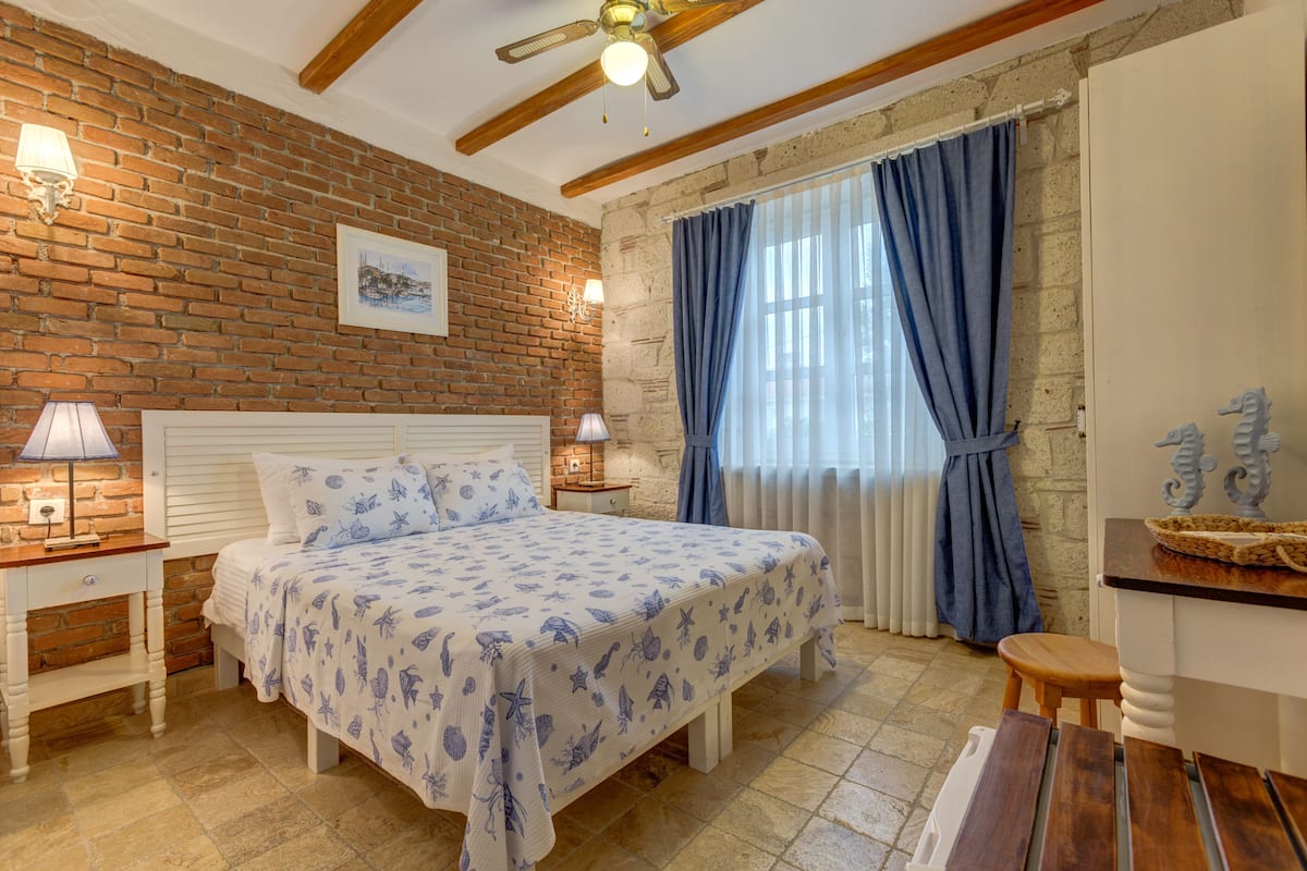 Un dormitorio con pared de ladrillo, una cama con colcha floral, dos lámparas de noche, un ventilador de techo de madera y una ventana con cortinas azules.