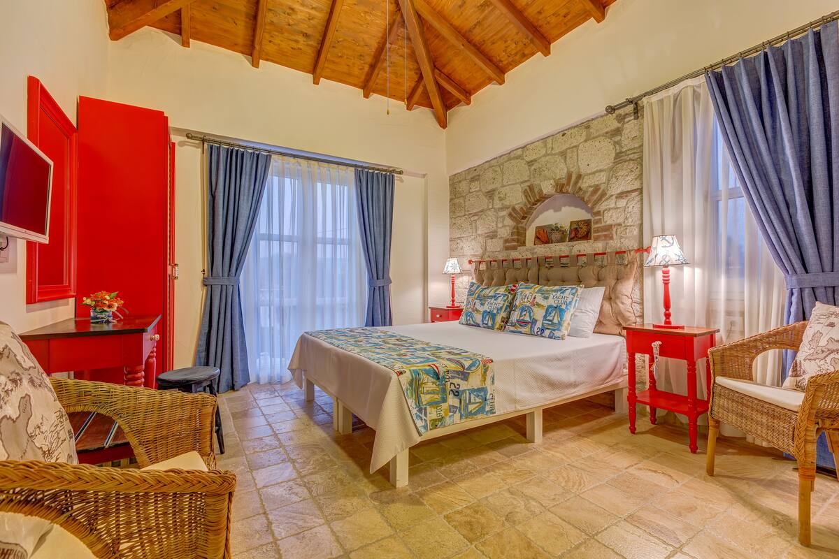 Un dormitorio con una cama, dos sillas de mimbre, una mesa roja, una pared de piedra y un televisor.
