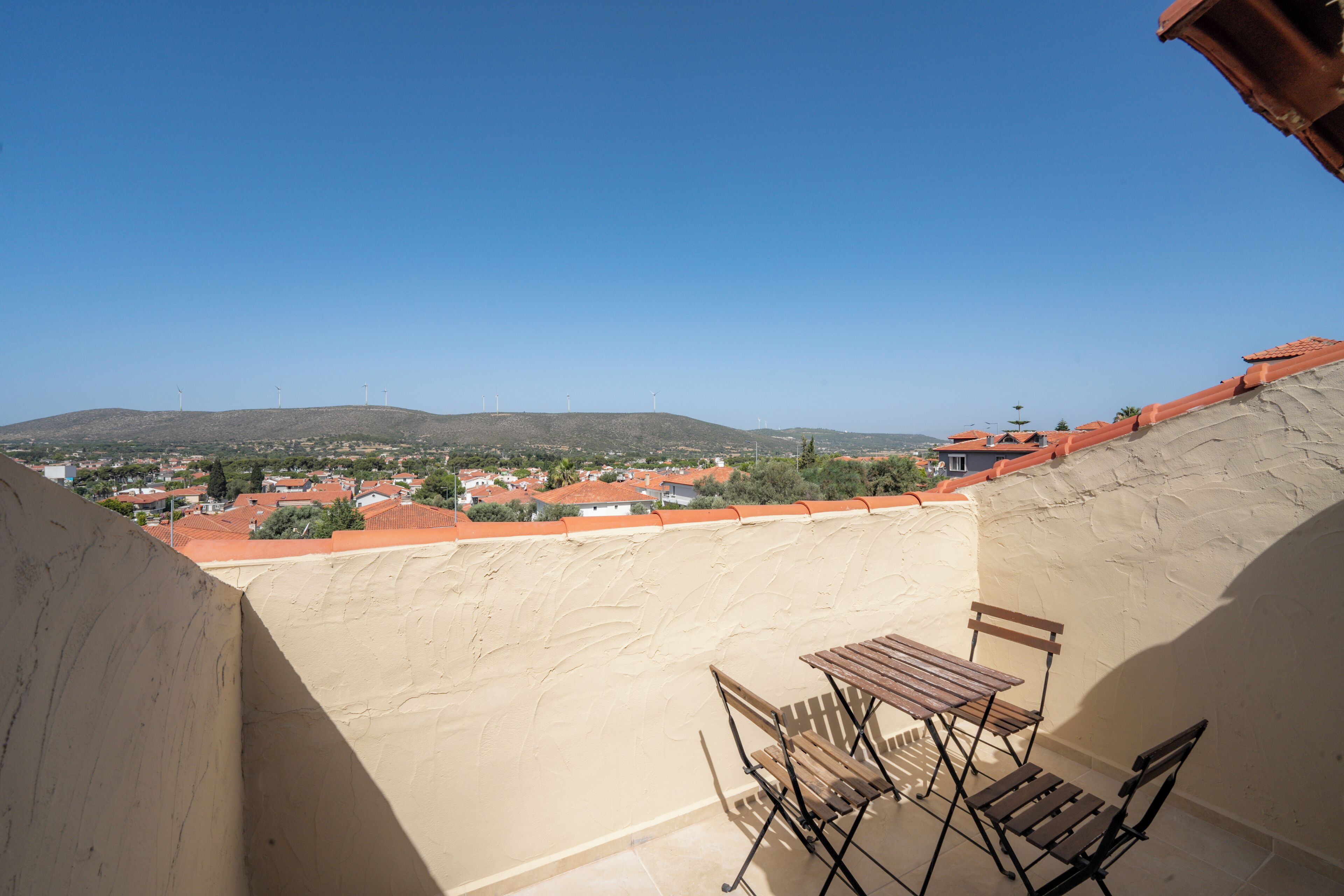 A balcony with a table and chairs, overlooking a town with red-roofed buildings and wind turbines in the distance.