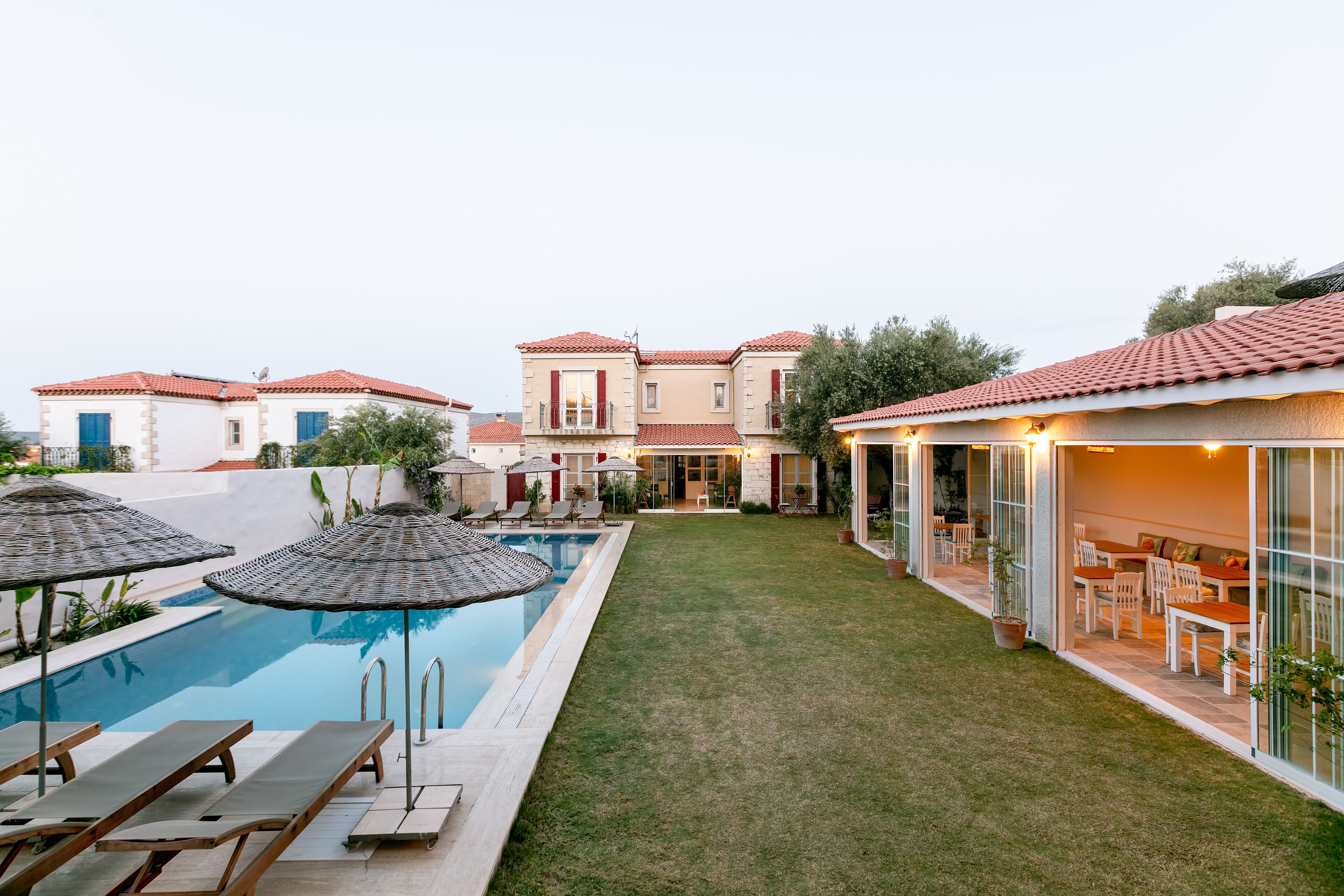 A pool area with lounge chairs and umbrellas, a grassy lawn, and a building with a red-tiled roof.