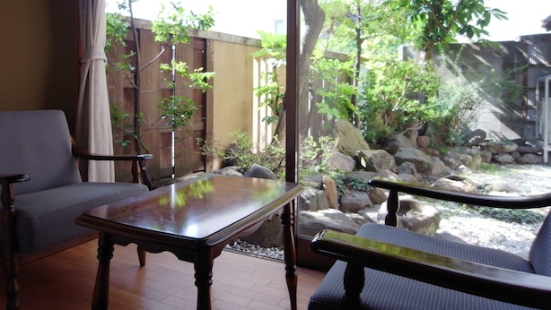 A room with a wooden table and two chairs, a view of a garden with rocks and trees, and a wooden fence. - Ryokan Yamamuro (Kanazawa)