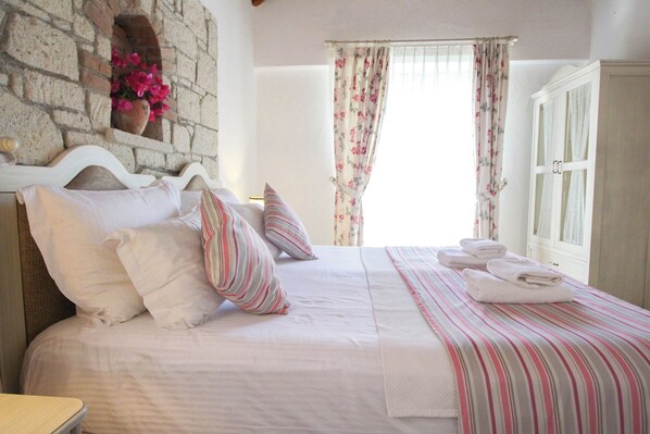 A neatly made bed with pillows and a striped blanket, a stone wall with a flower pot, and a window with floral curtains. - Alanarin Konak Alacati (Cesme)