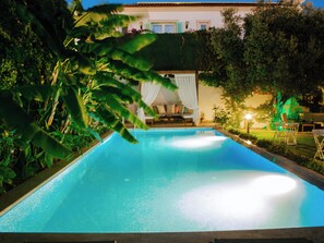 Una piscina con superficie de agua azul cristalina, rodeada de exuberante follaje y un edificio blanco con balcón al fondo.