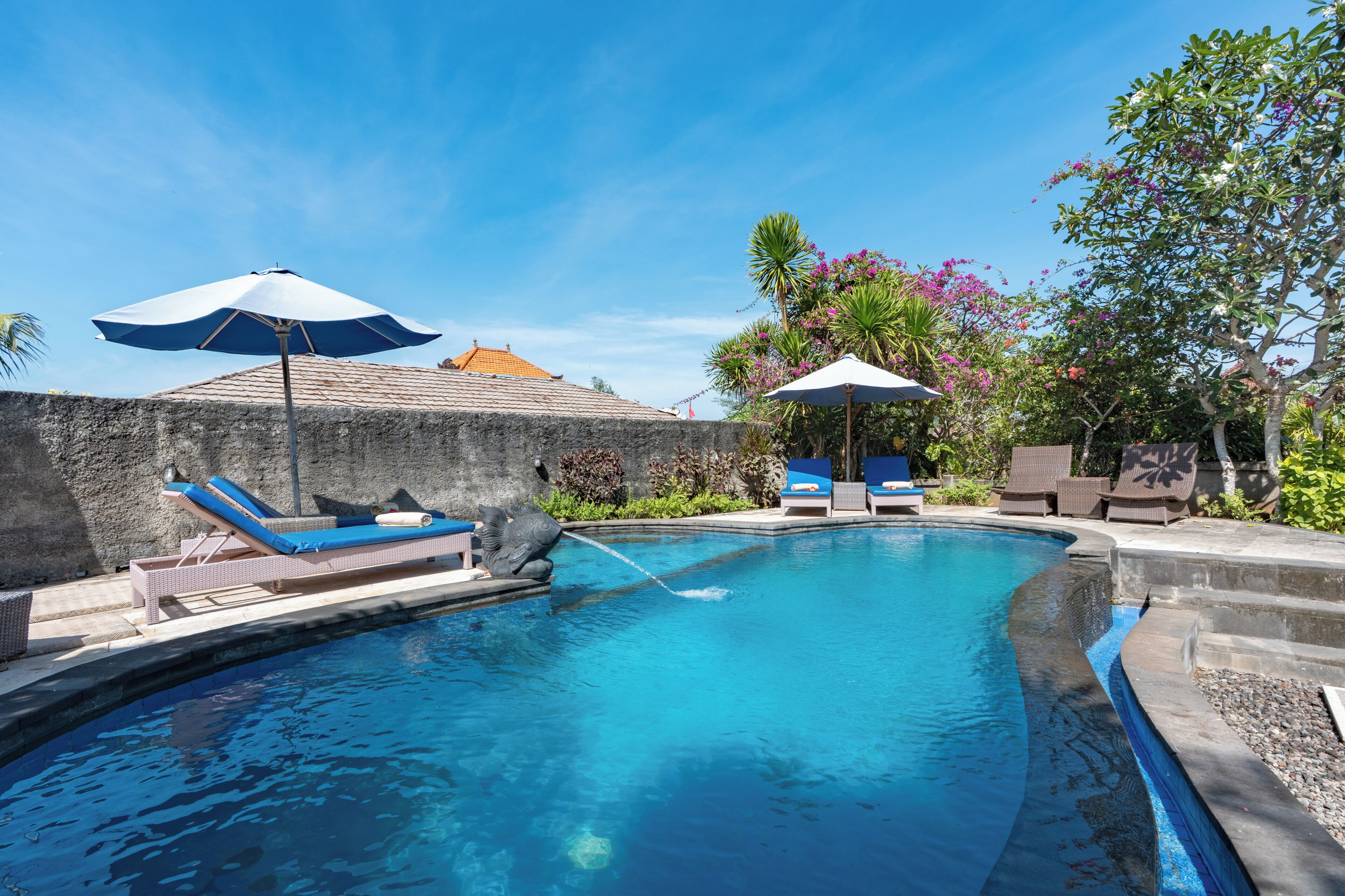 Una piscina con sillas de descanso y sombrillas, rodeada de vegetación y un muro de piedra.